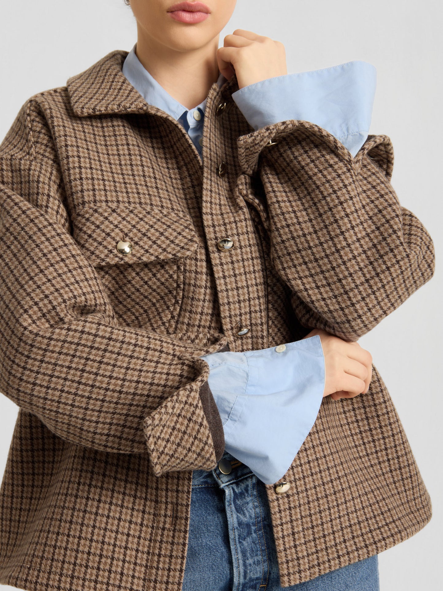 Eine Person, die die braune, karierte Jacke „Caitlin“ aus recycelter Wolle über einem hellblauen Hemd und blauen Jeans trägt, die Arme verschränkt und die Hand am Kinn. Ihr Gesicht ist teilweise zu sehen.