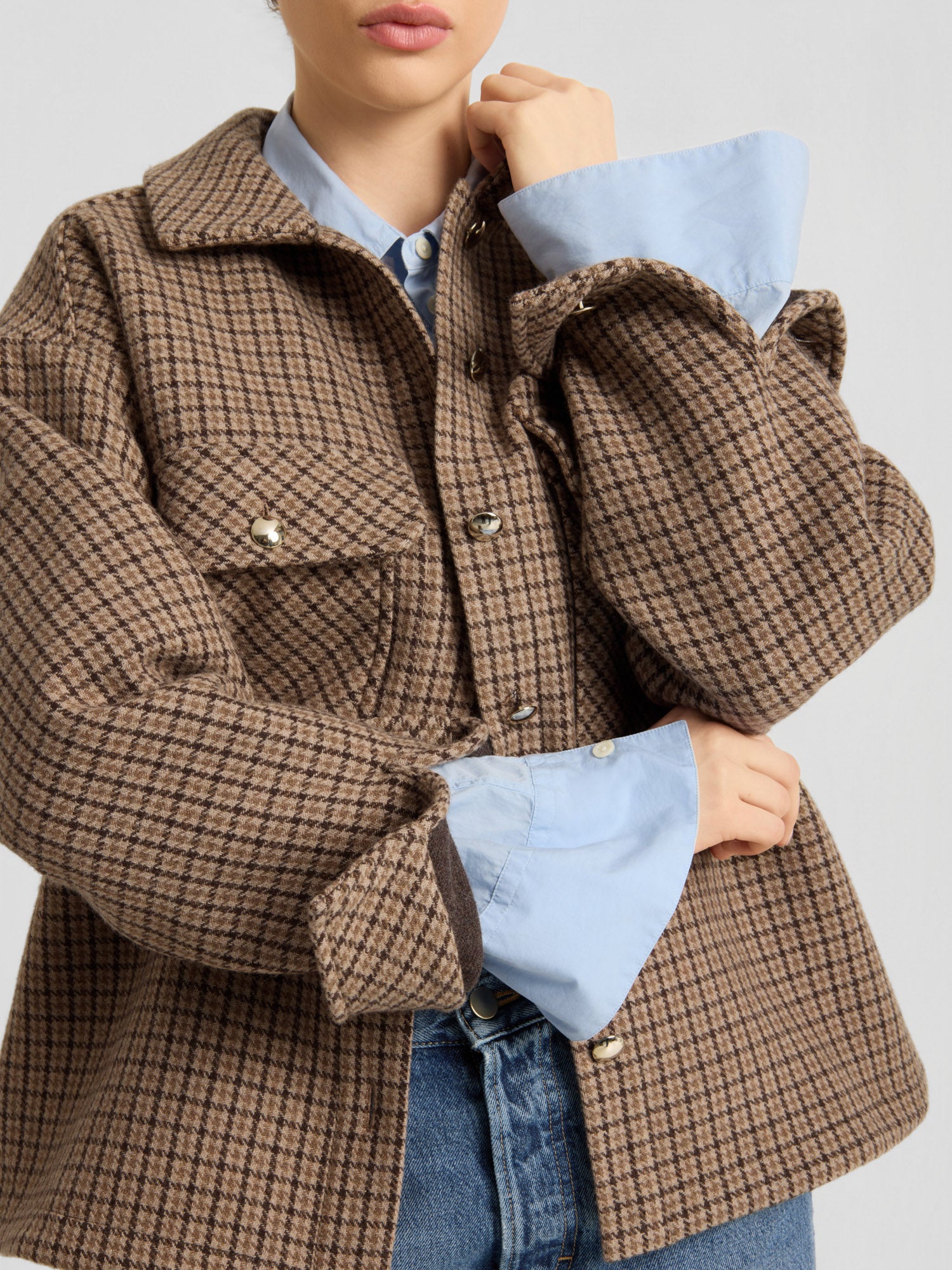 Eine Person, die die braune, karierte Jacke „Caitlin“ aus recycelter Wolle über einem hellblauen Hemd und blauen Jeans trägt, die Arme verschränkt und die Hand am Kinn. Ihr Gesicht ist teilweise zu sehen.