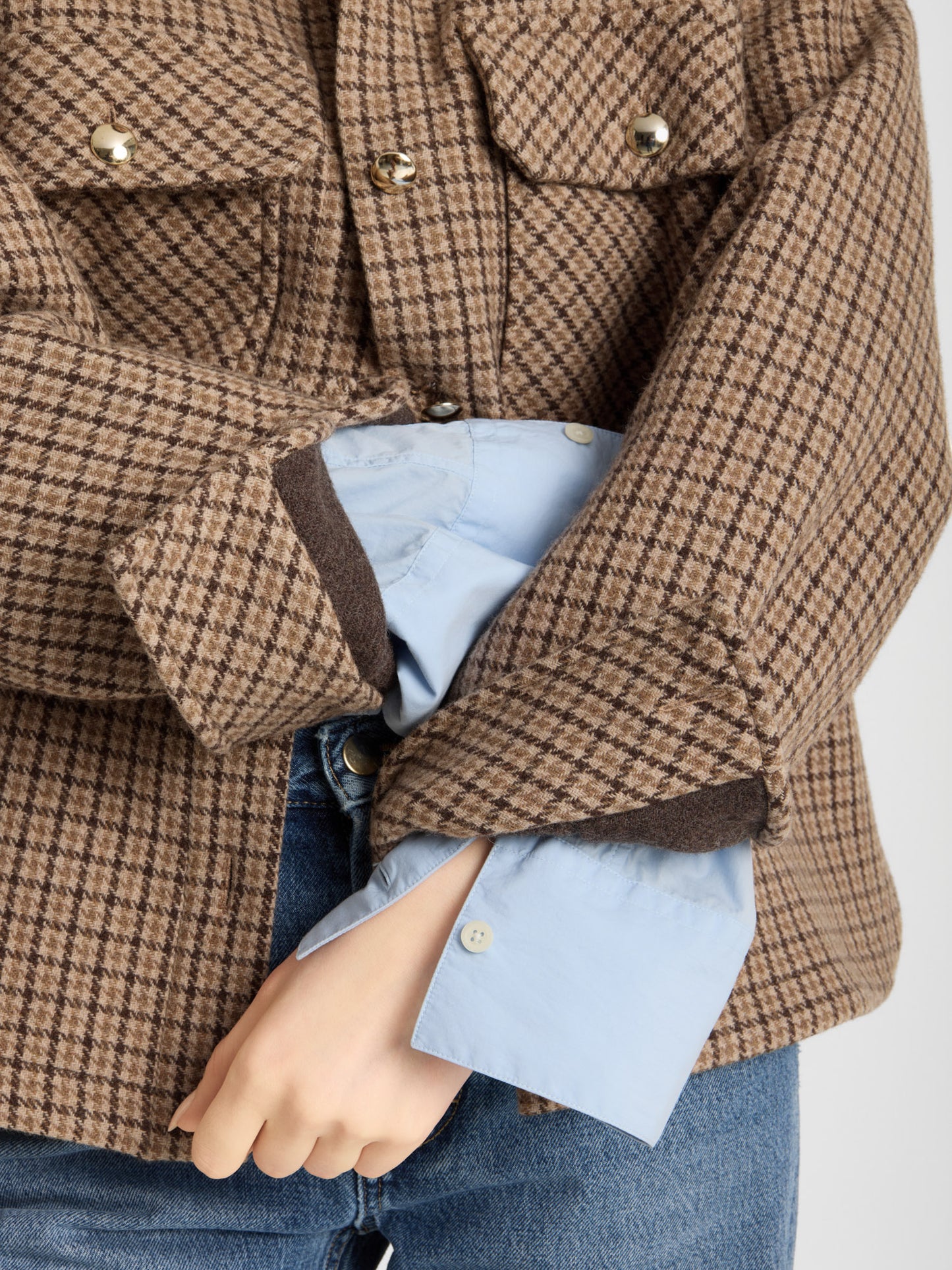 Eine Person trägt die braune, karierte Jacke „Caitlin“ aus recycelter Wolle über einem hellblauen Shirt und blauen Jeans und steht mit verschränkten Armen da, während eine Nahaufnahme die verschiedenen Lagen der Winterjacke sowie die Knopfdetails zeigt.