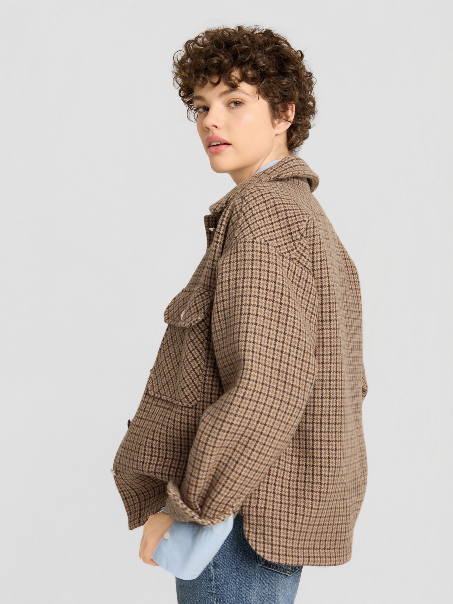 Eine Person mit kurzen Locken, die die braune Jacke „Caitlin“ über einem blauen Hemd und einer blauen Jeans trägt, steht seitlich und blickt vor einem schlichten hellgrauen Hintergrund in die Kamera.