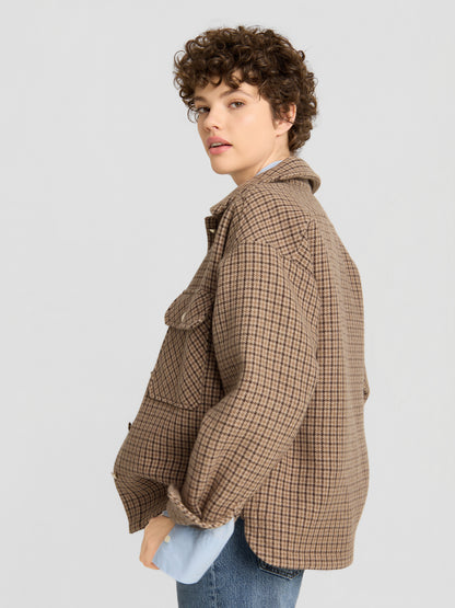 Eine Person mit kurzen Locken, die die braune Jacke „Caitlin“ über einem blauen Hemd und einer blauen Jeans trägt, steht seitlich und blickt vor einem schlichten hellgrauen Hintergrund in die Kamera.