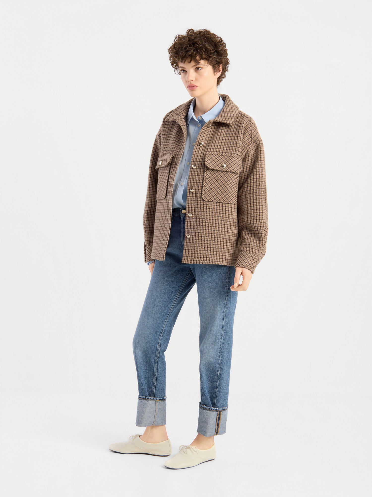 Eine Person mit kurzen Locken trägt die Jacke „Caitlin“, eine karierte braune Damen-Wolljacke aus recycelter Wolle, über einem blauen Hemd, dazu eine Jeans mit Umschlag und weiße Schuhe; sie steht vor einem schlichten weißen Hintergrund.