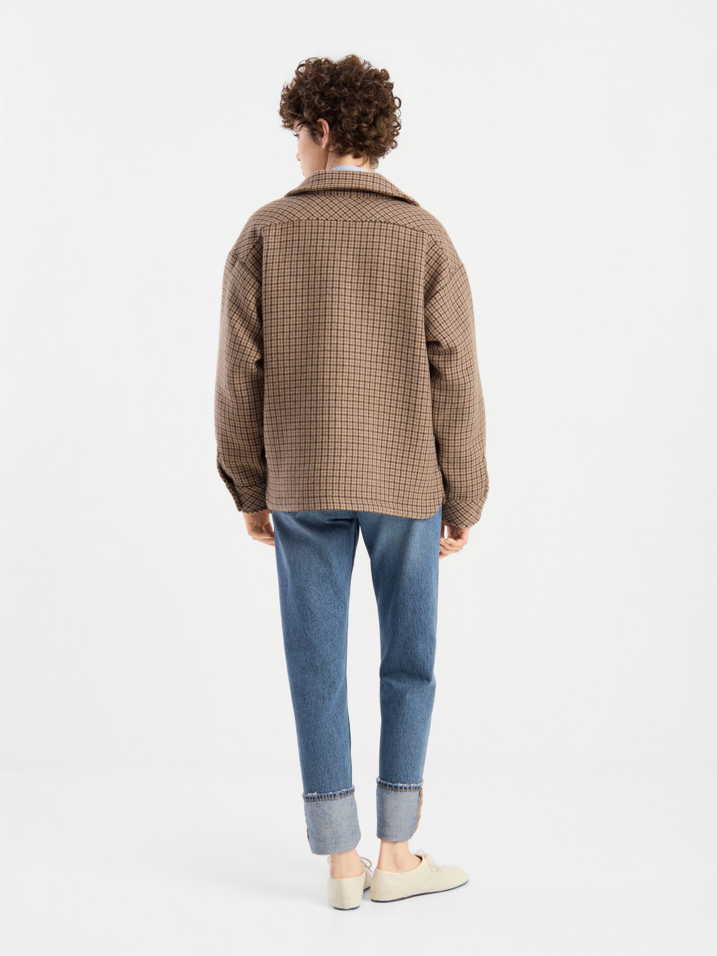 Eine Person mit lockigem Haar, die die braune karierte Jacke „Caitlin“ aus recycelter Wolle, eine blaue Jeans mit Umschlag und weiße Schuhe trägt, steht mit dem Rücken zur Kamera vor einem schlichten weißen Hintergrund.