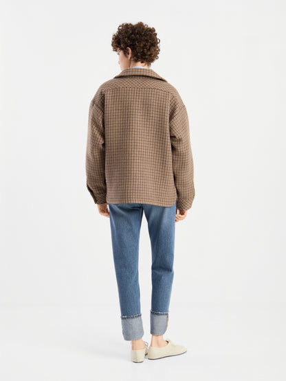 Eine Person mit lockigem Haar, die die braune karierte Jacke „Caitlin“ aus recycelter Wolle, eine blaue Jeans mit Umschlag und weiße Schuhe trägt, steht mit dem Rücken zur Kamera vor einem schlichten weißen Hintergrund.