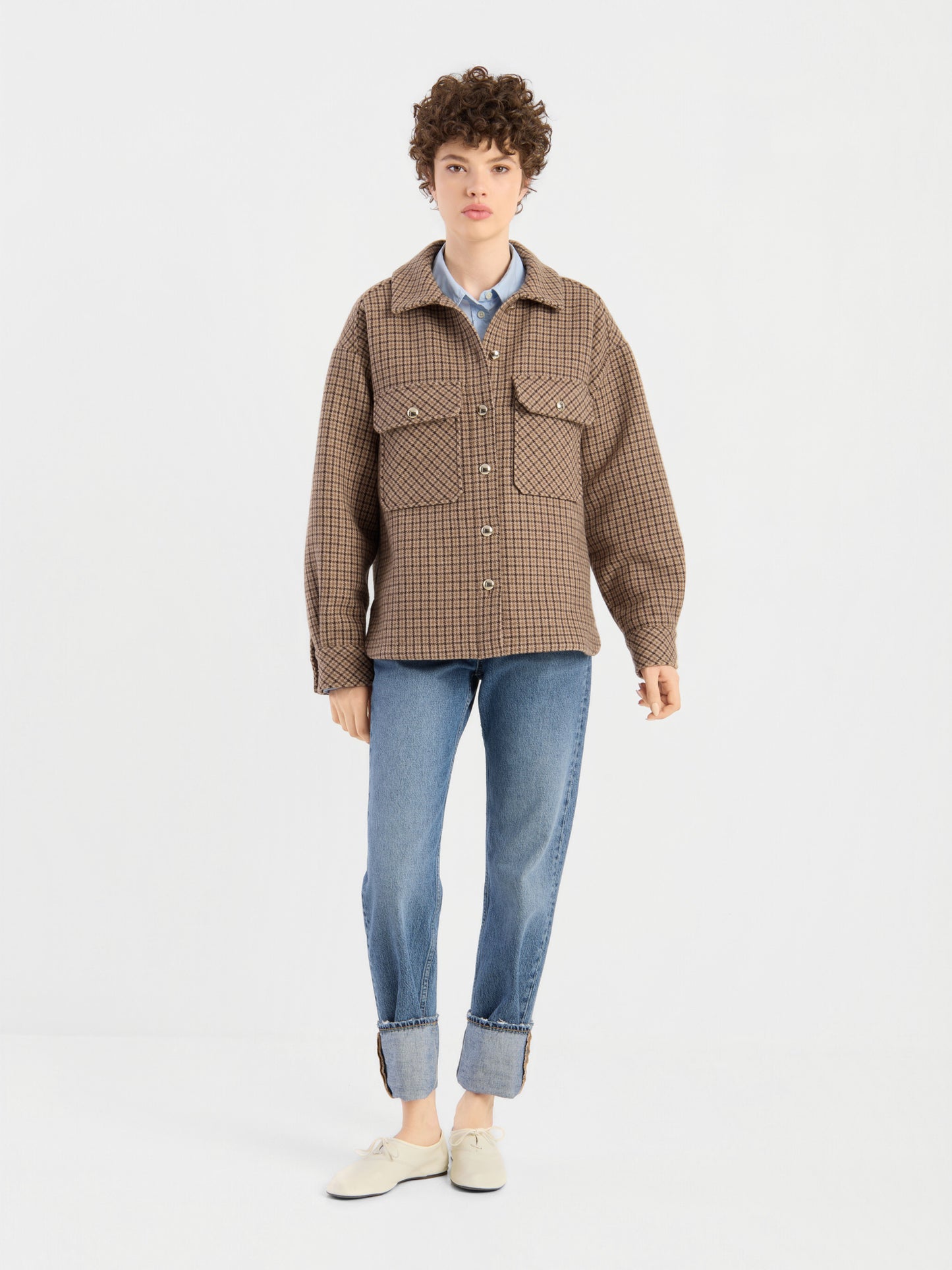 Eine Person mit kurzen Locken trägt die braun karierte Jacke „Caitlin“ aus recycelter Wolle über einem blauen Hemd, dazu eine blaue Jeans mit Umschlag und weiße Schuhe; sie steht vor einem schlichten weißen Hintergrund.