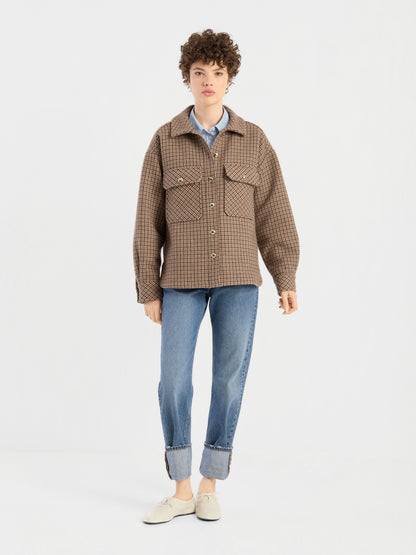 Eine Person mit kurzen Locken trägt die braun karierte Jacke „Caitlin“ aus recycelter Wolle über einem blauen Hemd, dazu eine blaue Jeans mit Umschlag und weiße Schuhe; sie steht vor einem schlichten weißen Hintergrund.