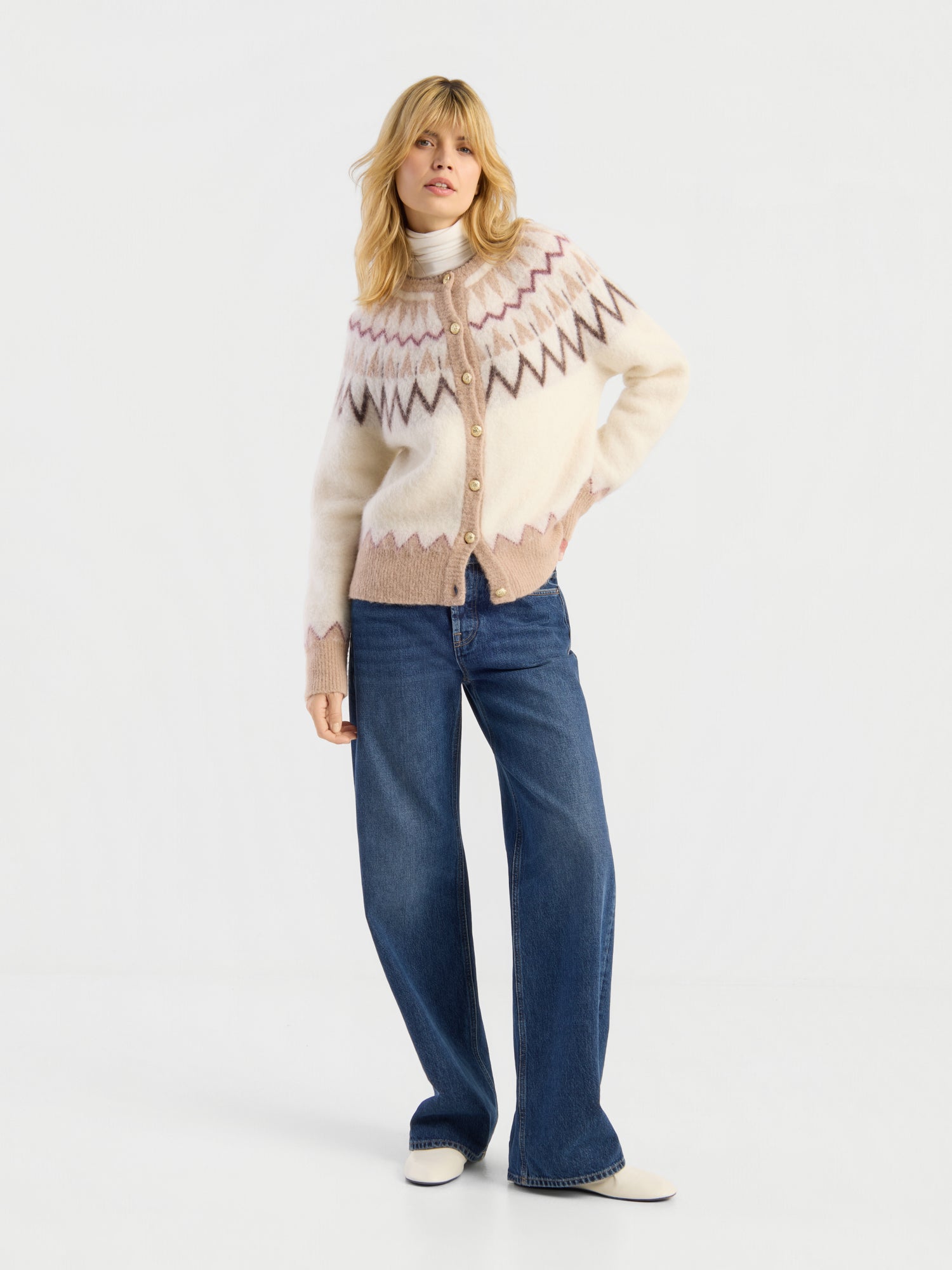 Eine Frau mit blonden Haaren posiert vor einem weißen Hintergrund in einer gemusterten Strickjacke, einem hellen Rollkragenpullover und einer NOLA Jeans Dark Blue – Wide Leg Damenjeans. Sie rundet ihren Look mit weißen Schuhen und einer lässigen Haltung ab, bei der sie die Hände in die Taschen gesteckt hat.