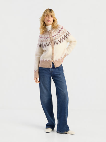 Eine Frau mit blonden Haaren posiert vor einem weißen Hintergrund in einer gemusterten Strickjacke, einem hellen Rollkragenpullover und einer NOLA Jeans Dark Blue – Wide Leg Damenjeans. Sie rundet ihren Look mit weißen Schuhen und einer lässigen Haltung ab, bei der sie die Hände in die Taschen gesteckt hat.