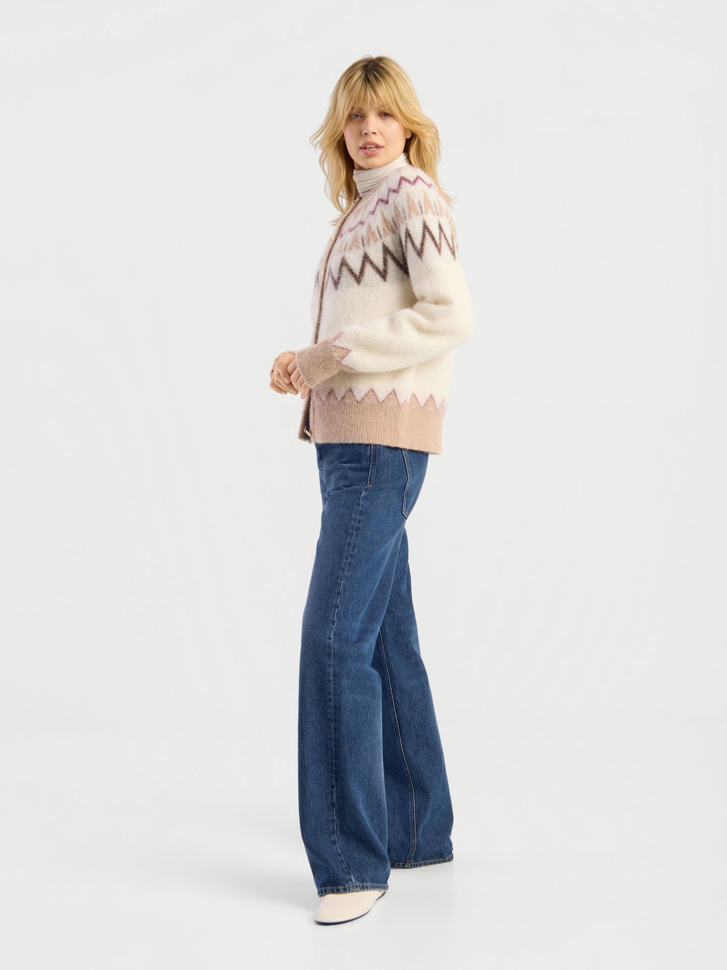 Eine Person, die die Jacke „Brushed Norweger“ in Offwhite-Beige, eine blaue Schlagjeans und weiße Schuhe trägt, steht in einer entspannten Haltung vor einem schlichten weißen Hintergrund.