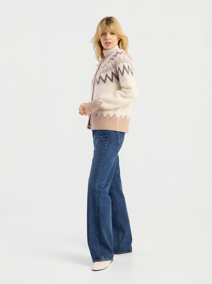 Eine Person, die die Jacke „Brushed Norweger“ in Offwhite-Beige, eine blaue Schlagjeans und weiße Schuhe trägt, steht in einer entspannten Haltung vor einem schlichten weißen Hintergrund.