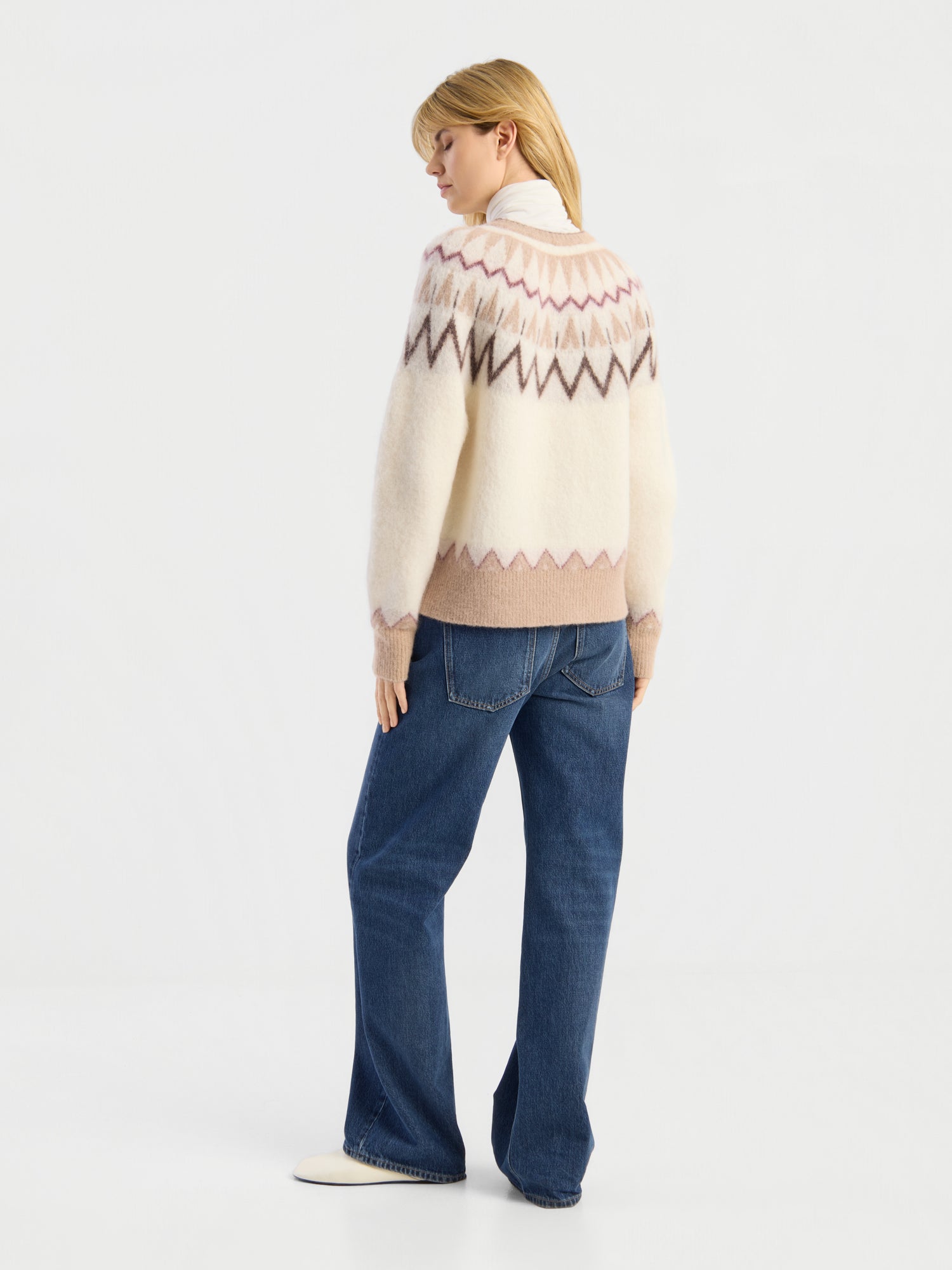 Eine Frau steht mit dem Rücken zur Kamera und trägt die „Brushed Norweger Jacket“ in Off-White-Beige, kombiniert mit blauen Jeans und weißen Schuhen, vor einem schlichten weißen Hintergrund.