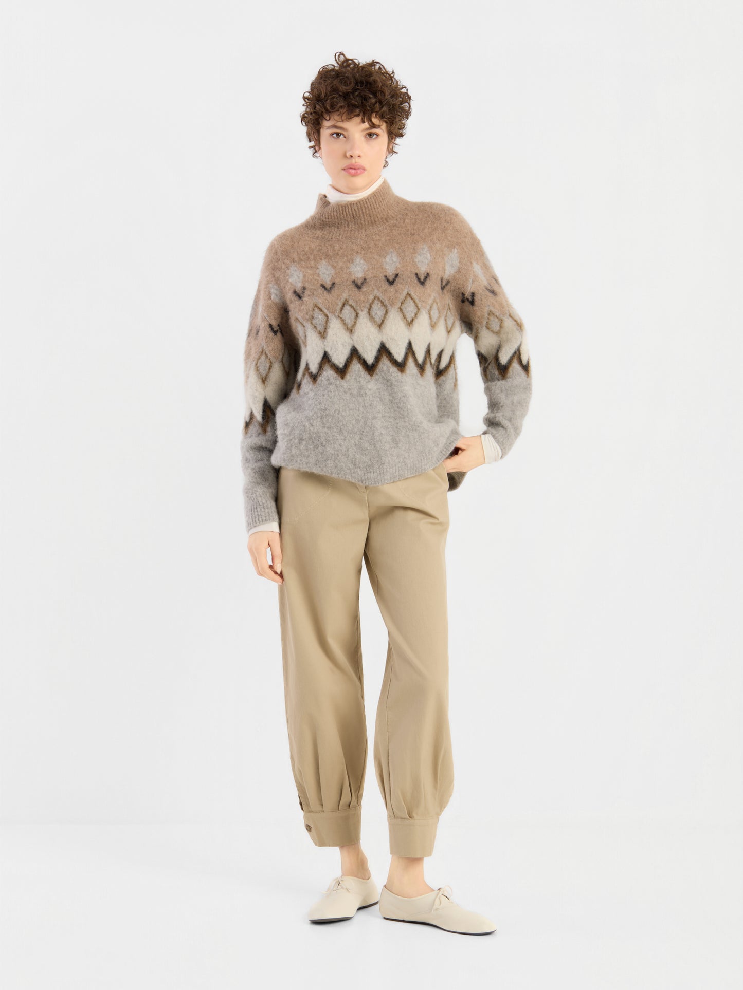 Eine Person steht vor einem weißen Hintergrund und trägt den „Brushed Norweger Sweater Beige“ aus luxuriösem Baby-Suri-Alpaka, eine hellbraune Hose mit Umschlag sowie beige Schuhe; sie hat kurzes, lockiges Haar und einen neutralen Gesichtsausdruck.