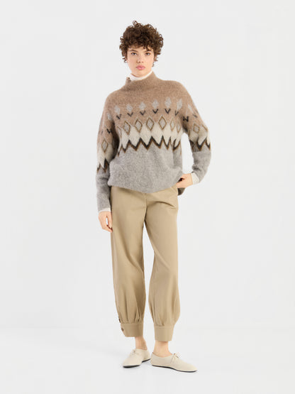 Eine Person steht vor einem weißen Hintergrund und trägt den „Brushed Norweger Sweater Beige“ aus luxuriösem Baby-Suri-Alpaka, eine hellbraune Hose mit Umschlag sowie beige Schuhe; sie hat kurzes, lockiges Haar und einen neutralen Gesichtsausdruck.