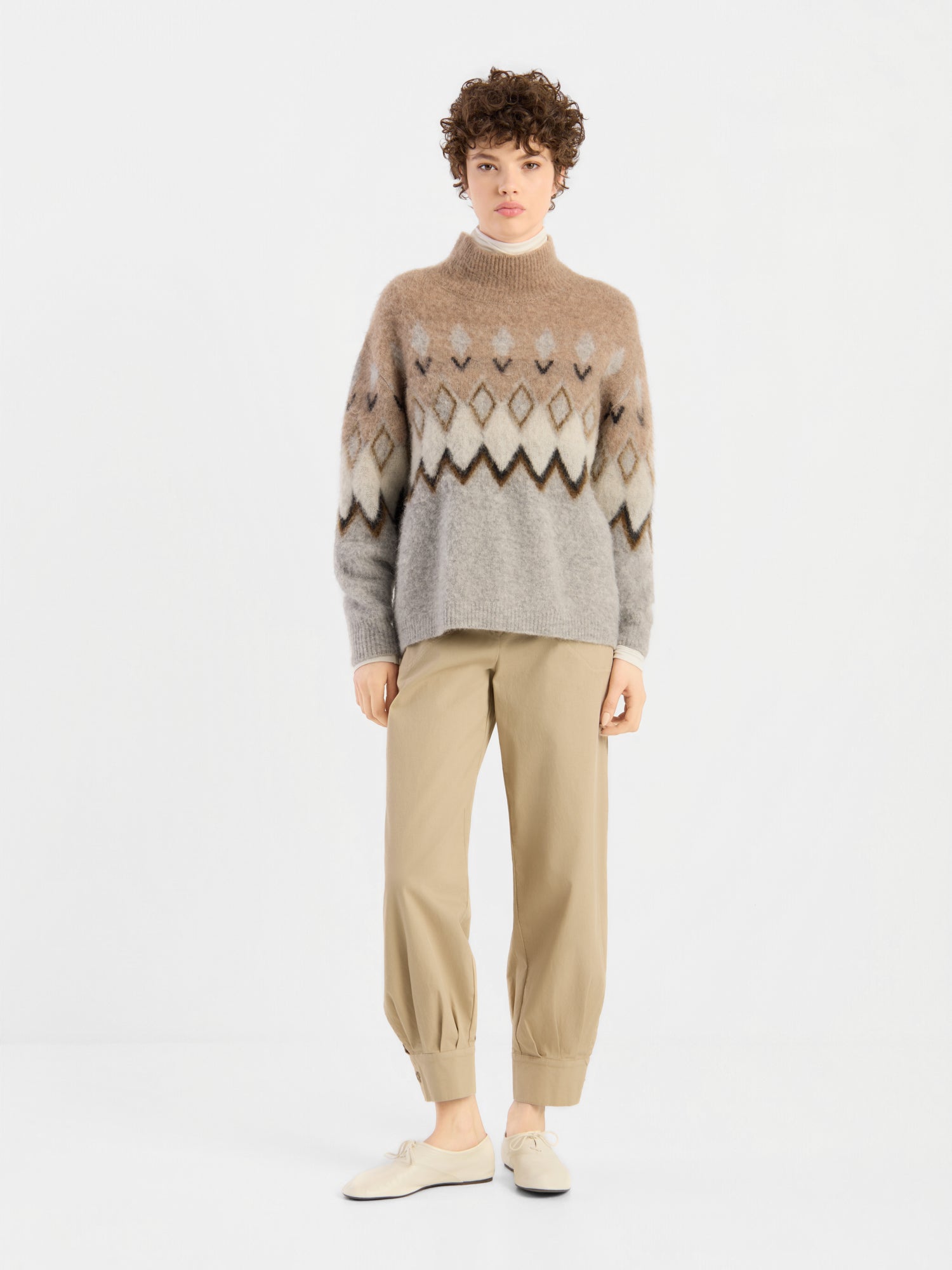 Eine Person steht vor einem schlichten weißen Hintergrund, blickt direkt in die Kamera und trägt den „Brushed Norweger Sweater Beige“ aus luxuriösem Baby-Suri-Alpaka, kombiniert mit einer khakifarbenen Hose und cremefarbenen Schuhen. Diese stilvolle Strickware wird stolz in Italien hergestellt.