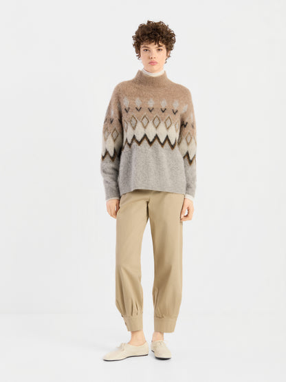Eine Person steht vor einem schlichten weißen Hintergrund, blickt direkt in die Kamera und trägt den „Brushed Norweger Sweater Beige“ aus luxuriösem Baby-Suri-Alpaka, kombiniert mit einer khakifarbenen Hose und cremefarbenen Schuhen. Diese stilvolle Strickware wird stolz in Italien hergestellt.