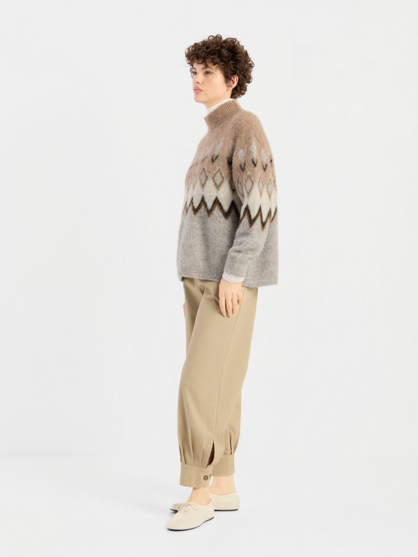 Eine Person mit kurzen Locken trägt den „Brushed Norweger Sweater Beige“ – einen in Italien hergestellten Pullover mit einem Muster aus Beige- und Brauntönen –, dazu eine hellbraune Hose mit Umschlag und weiße Slipper. Sie steht seitlich vor einem schlichten weißen Hintergrund.