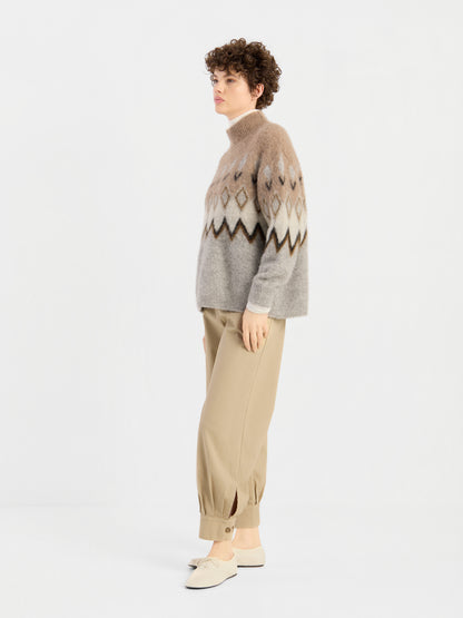 Eine Person mit kurzen Locken trägt den „Brushed Norweger Sweater Beige“ – einen in Italien hergestellten Pullover mit einem Muster aus Beige- und Brauntönen –, dazu eine hellbraune Hose mit Umschlag und weiße Slipper. Sie steht seitlich vor einem schlichten weißen Hintergrund.