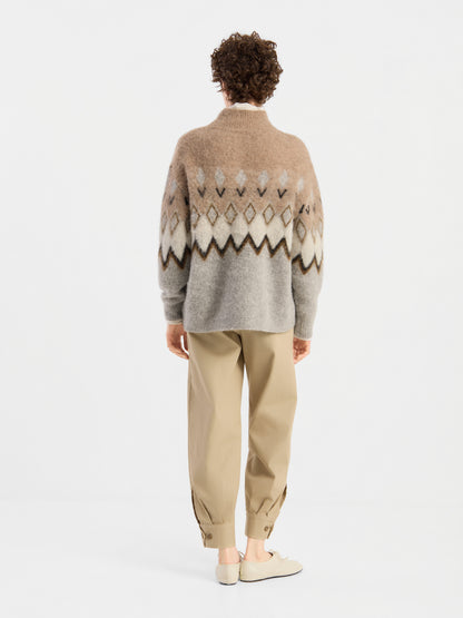 Eine Person steht mit dem Rücken zur Kamera und trägt einen beigen „Brushed Norweger“-Pullover, eine khakifarbene Hose mit Umschlag und helle Schuhe vor einem schlichten weißen Hintergrund.