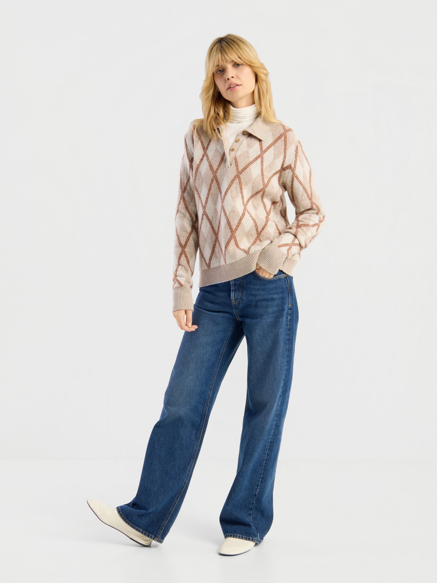 Eine Frau steht vor einem schlichten weißen Hintergrund und trägt den Pullover „Polo Diamond“ über einem weißen Oberteil, eine blaue Jeans mit weitem Bein und weiße Schuhe. Sie hat blondes Haar und posiert lässig mit einer Hand in der Hosentasche.