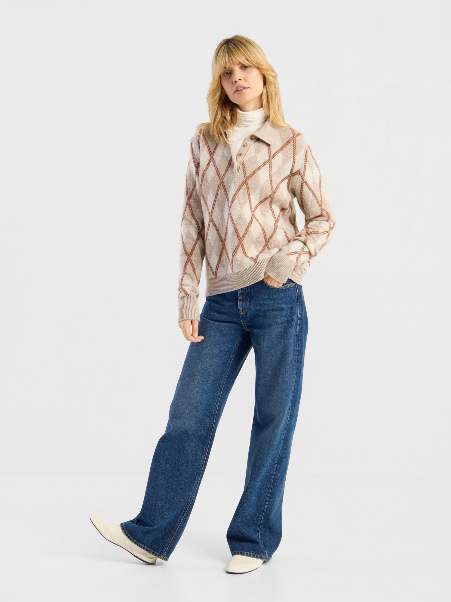 Eine Frau steht vor einem schlichten weißen Hintergrund und trägt den Pullover „Polo Diamond“ über einem weißen Oberteil, eine blaue Jeans mit weitem Bein und weiße Schuhe. Sie hat blondes Haar und posiert lässig mit einer Hand in der Hosentasche.