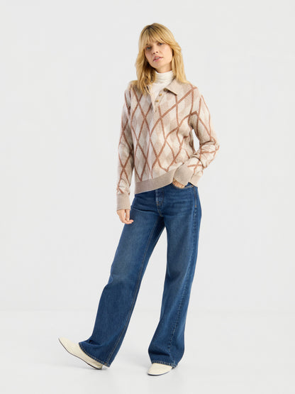 Eine Frau steht vor einem schlichten weißen Hintergrund und trägt den Pullover „Polo Diamond“ über einem weißen Oberteil, eine blaue Jeans mit weitem Bein und weiße Schuhe. Sie hat blondes Haar und posiert lässig mit einer Hand in der Hosentasche.