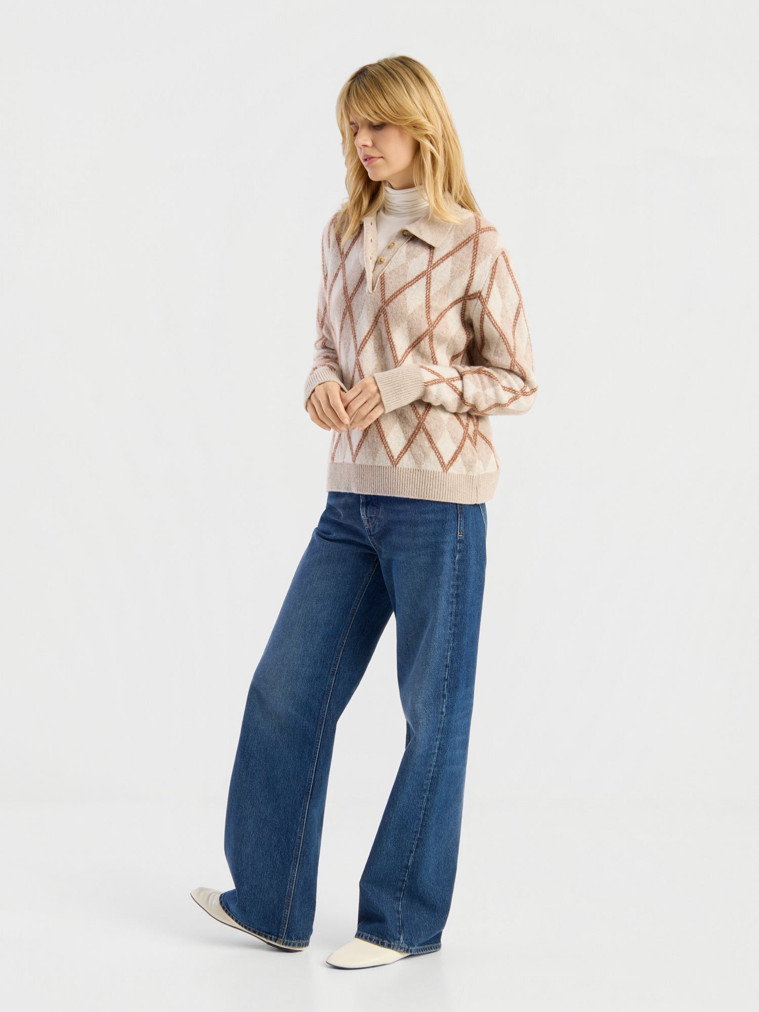 Vor einem schlichten Hintergrund steht eine Frau, die den „Pullover Polo Diamond“ trägt – einen beige-rosa Argyle-Pullover über einem weißen Rollkragenpullover –, dazu blaue Jeans mit weitem Bein und weiße Schuhe. Sie blickt leicht zur Seite, wobei ein Fuß nach vorne gestellt ist.
