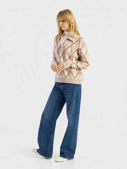 Vor einem schlichten Hintergrund steht eine Frau, die den „Pullover Polo Diamond“ trägt – einen beige-rosa Argyle-Pullover über einem weißen Rollkragenpullover –, dazu blaue Jeans mit weitem Bein und weiße Schuhe. Sie blickt leicht zur Seite, wobei ein Fuß nach vorne gestellt ist.
