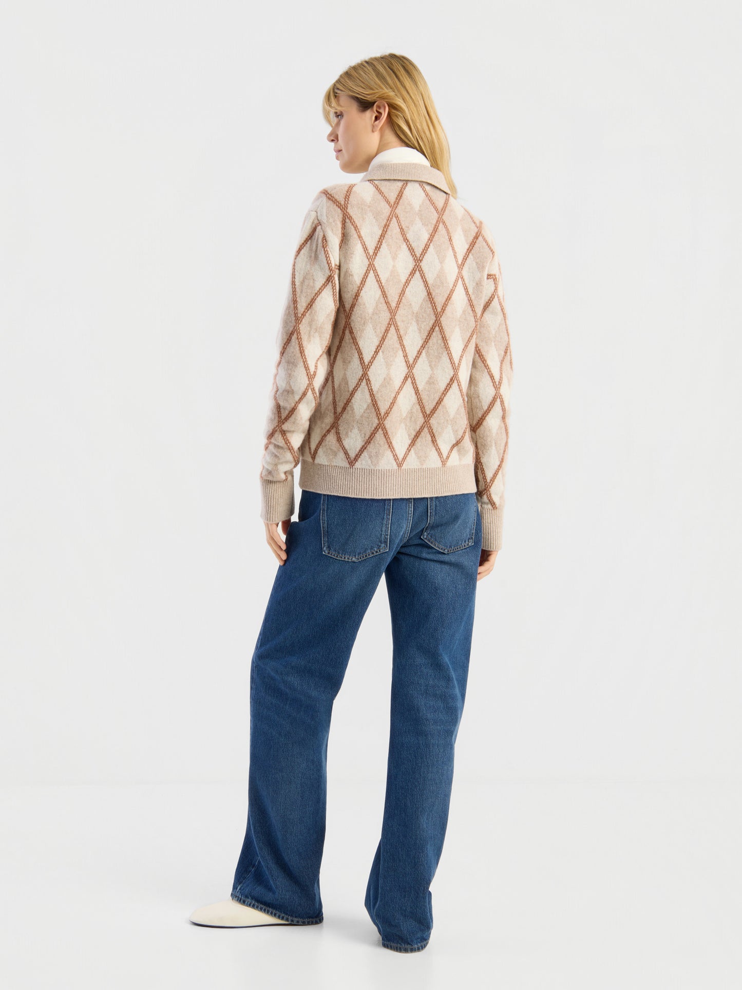 Vor einem schlichten weißen Hintergrund ist eine Person von hinten zu sehen, die den Pullover „Polo Diamond“ in Beige trägt, dazu blaue Jeans und weiße Schuhe.