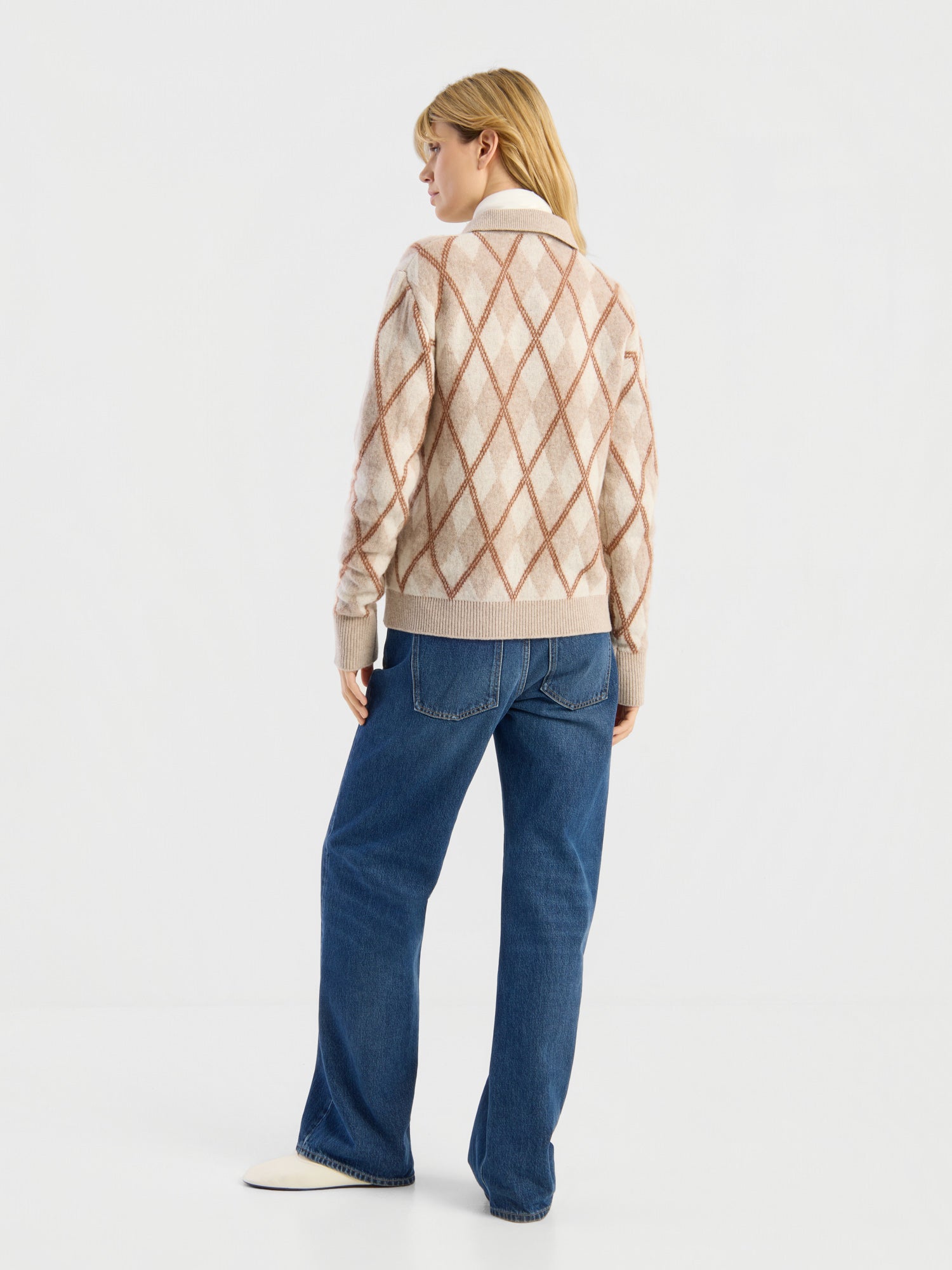 Vor einem schlichten weißen Hintergrund ist eine Person von hinten zu sehen, die den Pullover „Polo Diamond“ in Beige trägt, dazu blaue Jeans und weiße Schuhe.