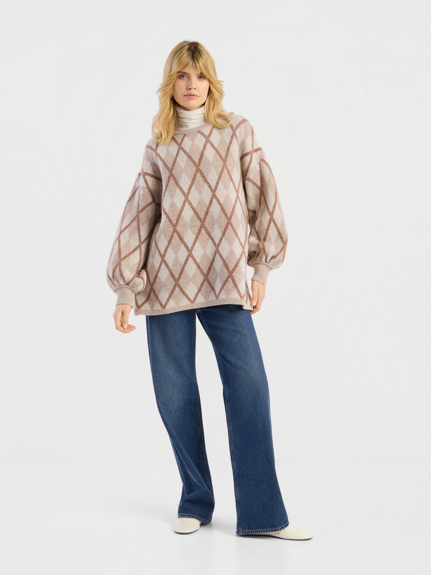 Eine Person, die den Pullover „Antonia Diamond“ trägt – einen beigen Pullover mit Rautenmuster und Ballonärmeln –, kombiniert mit einem hellen Rollkragenpullover, einer blau-weit geschnittenen Jeans und weißen Schuhen, steht vor einem schlichten weißen Hintergrund.