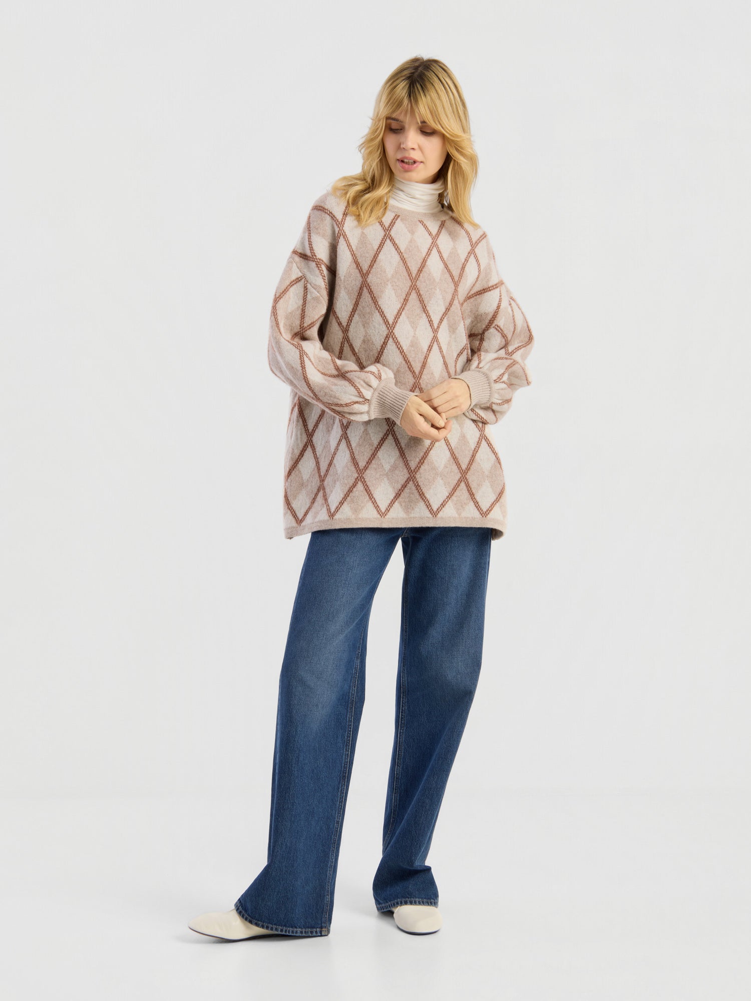Eine Person, die den Pullover „Antonia Diamond“ mit einem cremefarbenen und braunen Argyle-Muster trägt, kombiniert mit einer blau-weit geschnittenen Jeans und weißen Schuhen, steht vor einem schlichten weißen Hintergrund.
