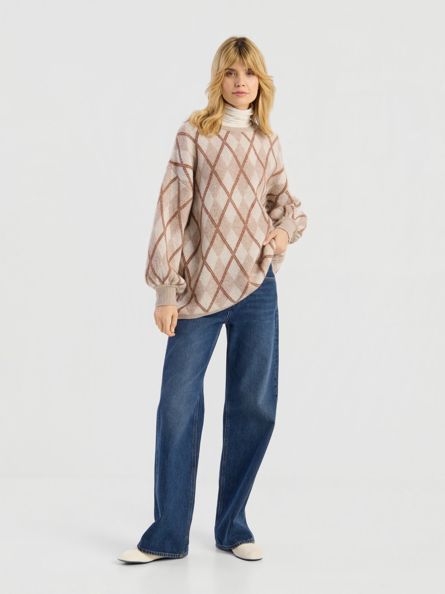 Dieser Look, bestehend aus dem Pullover „Antonia Diamond“ – einem beige-braun karierten Oversize-Damenpullover aus Merinowolle –, der über einem weißen Rollkragenpullover getragen wird, dazu eine blaue Hose mit weitem Bein und weiße Schuhe, hebt sich deutlich von einem schlichten, hellen Hintergrund ab. Der in Italien gefertigte, kuschelige Pullover vereint lässigen Stil mit erstklassiger Qualität.
