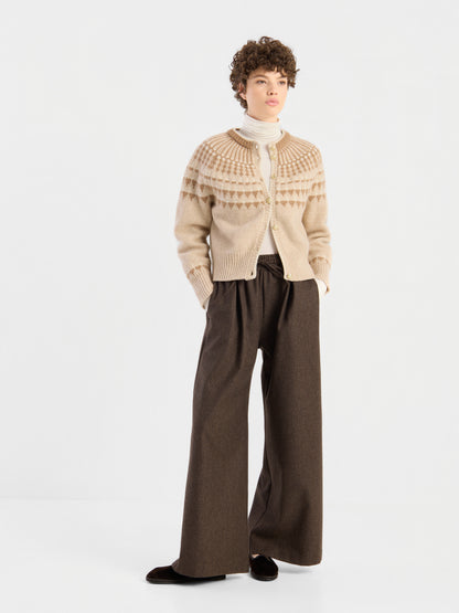 Die Person trägt die „Norweger Jacket Vol. 3“, eine beige gemusterte Strickjacke, darüber einen weißen Rollkragenpullover und eine weite braune Hose, und steht vor einem schlichten weißen Hintergrund.