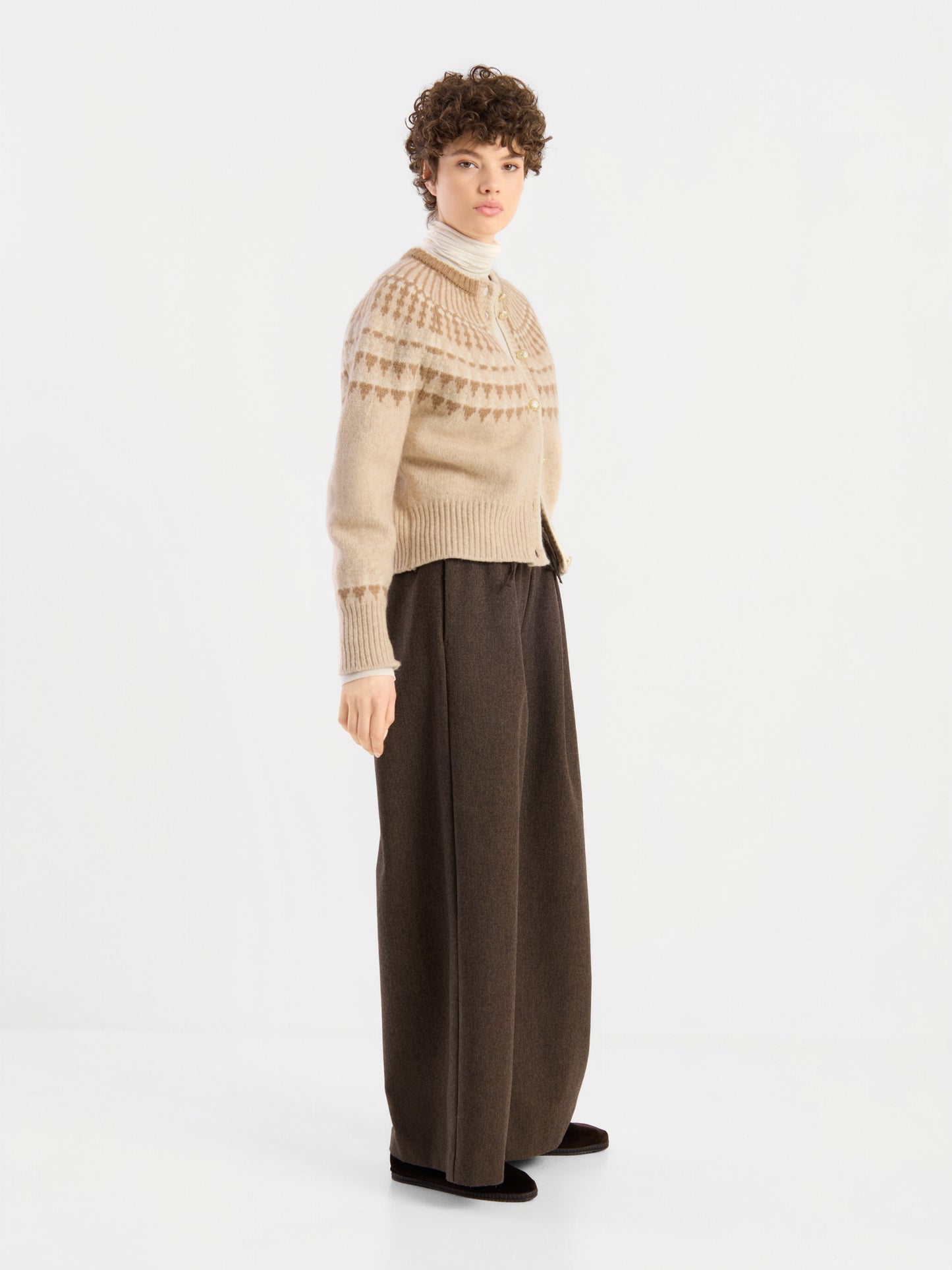 Eine Person, die die „Norweger Jacket Vol. 3“ – einen beige gemusterten Pullover – trägt, kombiniert mit einem weißen Rollkragenpullover und einer braunen Hose mit weitem Bein, steht vor einem schlichten weißen Hintergrund und blickt leicht zur Seite.