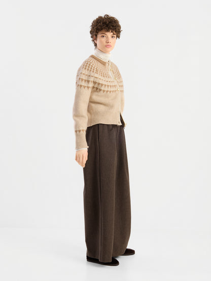 Eine Person, die die „Norweger Jacket Vol. 3“ – einen beige gemusterten Pullover – trägt, kombiniert mit einem weißen Rollkragenpullover und einer braunen Hose mit weitem Bein, steht vor einem schlichten weißen Hintergrund und blickt leicht zur Seite.