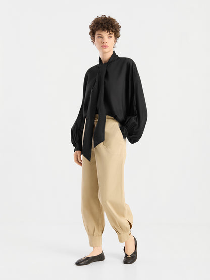 Eine Person, die eine locker sitzende schwarze Bluse „Antonia Bow Black“ mit einer großen Schleife, eine beige Hose mit Umschlag und schwarze flache Schuhe trägt und vor einem schlichten weißen Hintergrund steht.