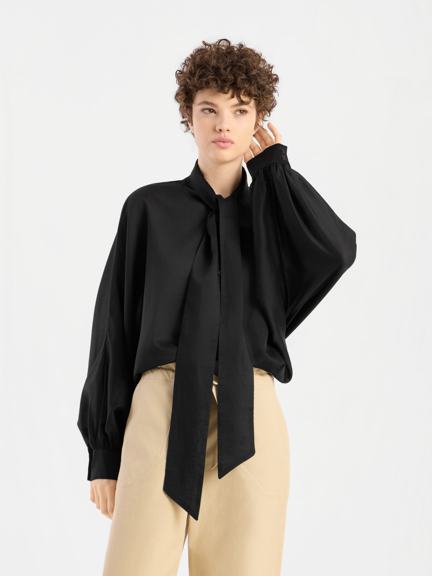 Eine Person mit kurzen Locken trägt eine locker sitzende Bluse „Antonia Bow Black“ mit einer langen Krawatte und einer beigen Hose mit hoher Taille und steht vor einem schlichten weißen Hintergrund.