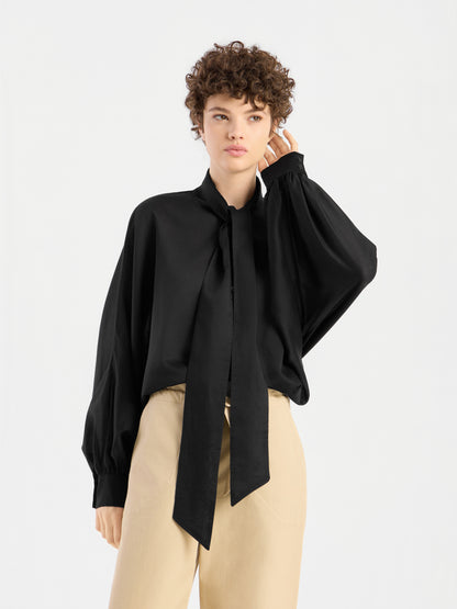 Eine Person mit kurzen Locken trägt eine locker sitzende Bluse „Antonia Bow Black“ mit einer langen Krawatte und einer beigen Hose mit hoher Taille und steht vor einem schlichten weißen Hintergrund.