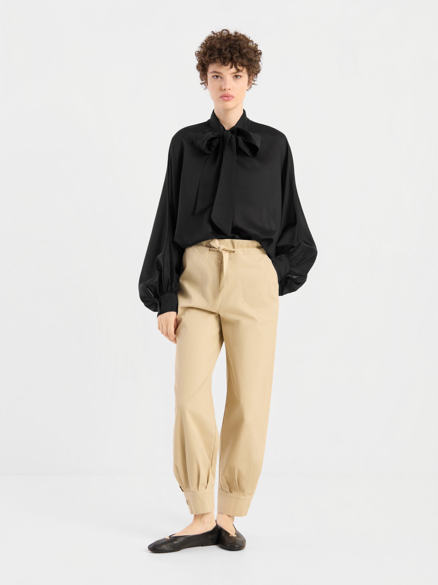 Eine Person steht vor einem schlichten weißen Hintergrund und trägt eine schwarze Bluse „Antonia Bow Black“ mit einer großen Schleife am Halsausschnitt, eine beige Hose mit hoher Taille und umgeschlagenen Säumen sowie schwarze Ballerinas.