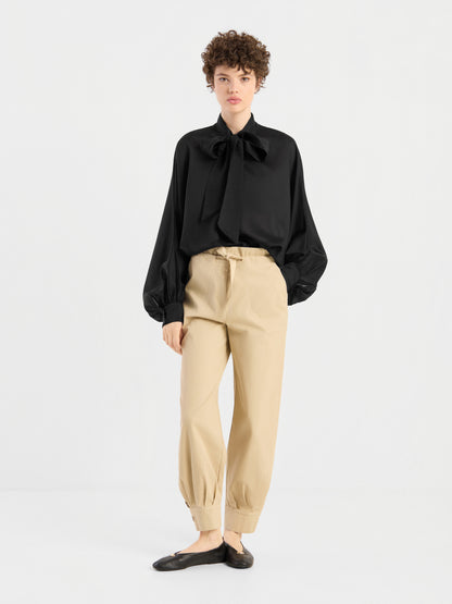 Eine Person steht vor einem schlichten weißen Hintergrund und trägt eine schwarze Bluse „Antonia Bow Black“ mit einer großen Schleife am Halsausschnitt, eine beige Hose mit hoher Taille und umgeschlagenen Säumen sowie schwarze Ballerinas.