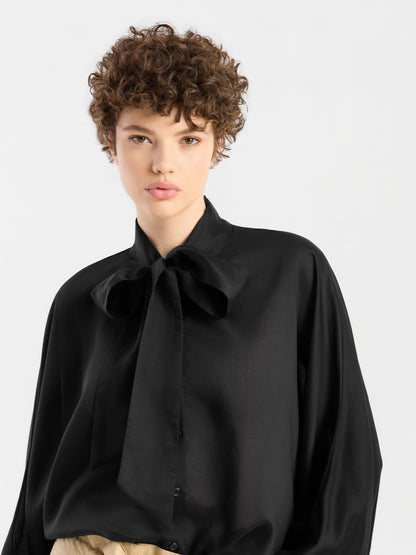 Eine Person mit kurzen Locken trägt die elegante Bluse „Antonia Bow Black“ mit einer großen Schleife am Kragen, dazu eine beige Hose, und steht vor einem schlichten, hellen Hintergrund.