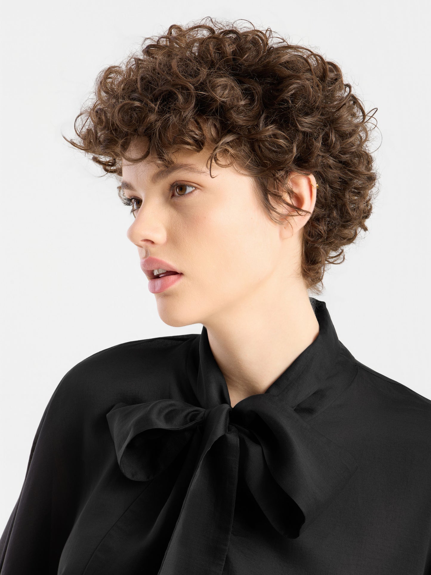 Eine Person mit kurzen, lockigen braunen Haaren blickt zur Seite und trägt vor einem schlichten weißen Hintergrund die „Bluse Antonia Bow Black“ – eine elegante schwarze Bluse mit einer großen Schleife am Kragen.