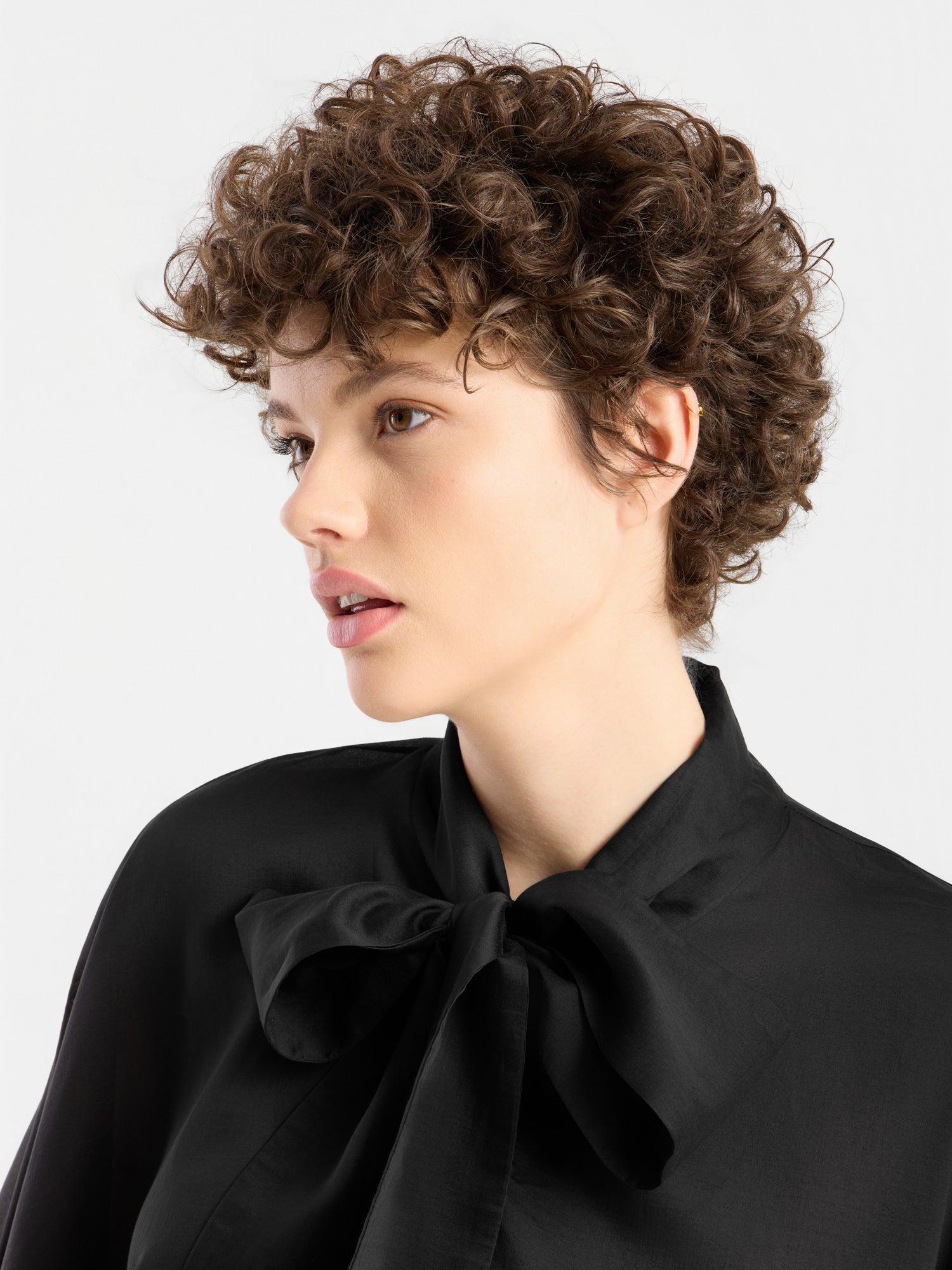 Eine Person mit kurzen, lockigen braunen Haaren blickt zur Seite und trägt vor einem schlichten weißen Hintergrund die „Bluse Antonia Bow Black“ – eine elegante schwarze Bluse mit einer großen Schleife am Kragen.
