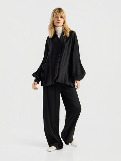 Eine Person, die die Hose „Aria Black“ als Teil eines locker sitzenden Outfits aus schwarzem Satin trägt, dazu ein passendes Hemd über einem weißen Rollkragenpullover und weiße Schuhe, steht vor einem schlichten, hellen Hintergrund.
