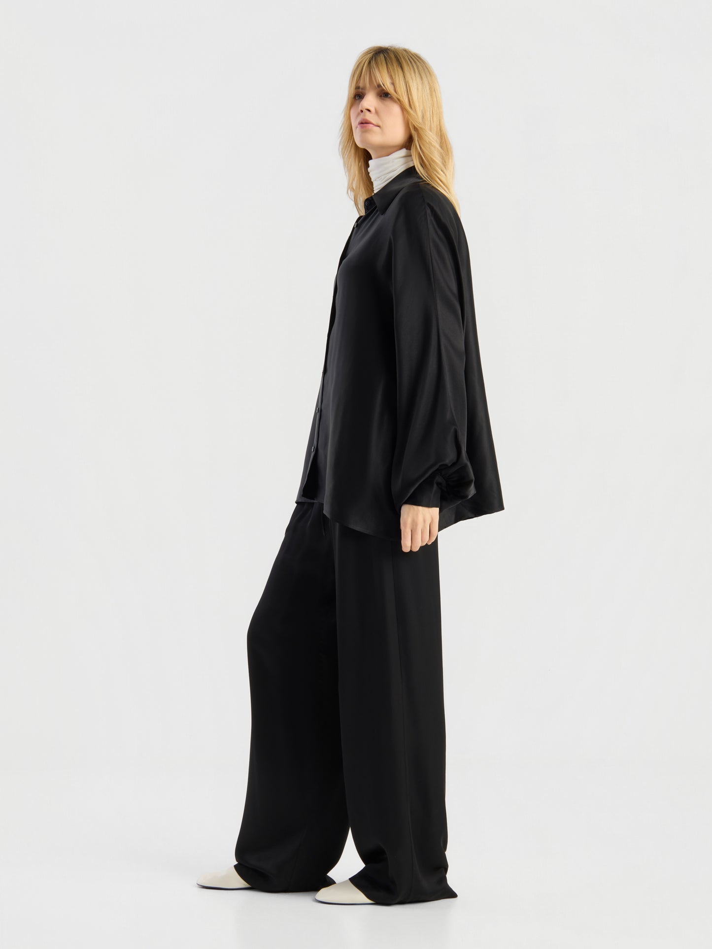 Eine Person steht seitlich vor einem schlichten weißen Hintergrund und trägt die Bluse „Antonia Shiny Black One Size“, die als lockeres schwarzes Hemd gestylt ist, dazu eine schwarze Hose mit weitem Bein, einen weißen Rollkragenpullover und weiße Schuhe.