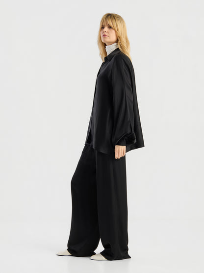 Eine Person steht seitlich vor einem schlichten weißen Hintergrund und trägt die Bluse „Antonia Shiny Black One Size“, die als lockeres schwarzes Hemd gestylt ist, dazu eine schwarze Hose mit weitem Bein, einen weißen Rollkragenpullover und weiße Schuhe.