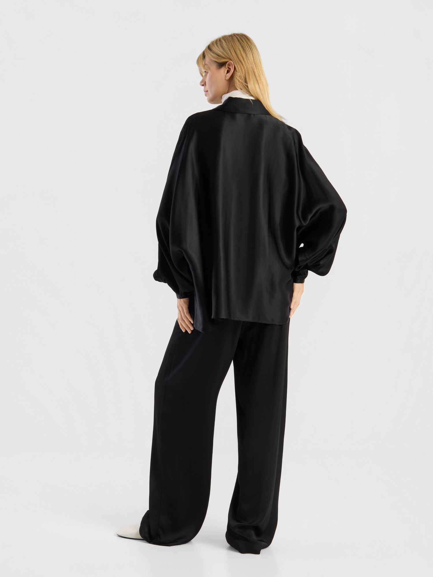 Eine Person mit langen blonden Haaren, die die Bluse „Antonia Shiny Black One Size“ in Kombination mit einer passenden Hose mit weitem Bein trägt, steht mit dem Rücken zur Kamera vor einem schlichten weißen Hintergrund.