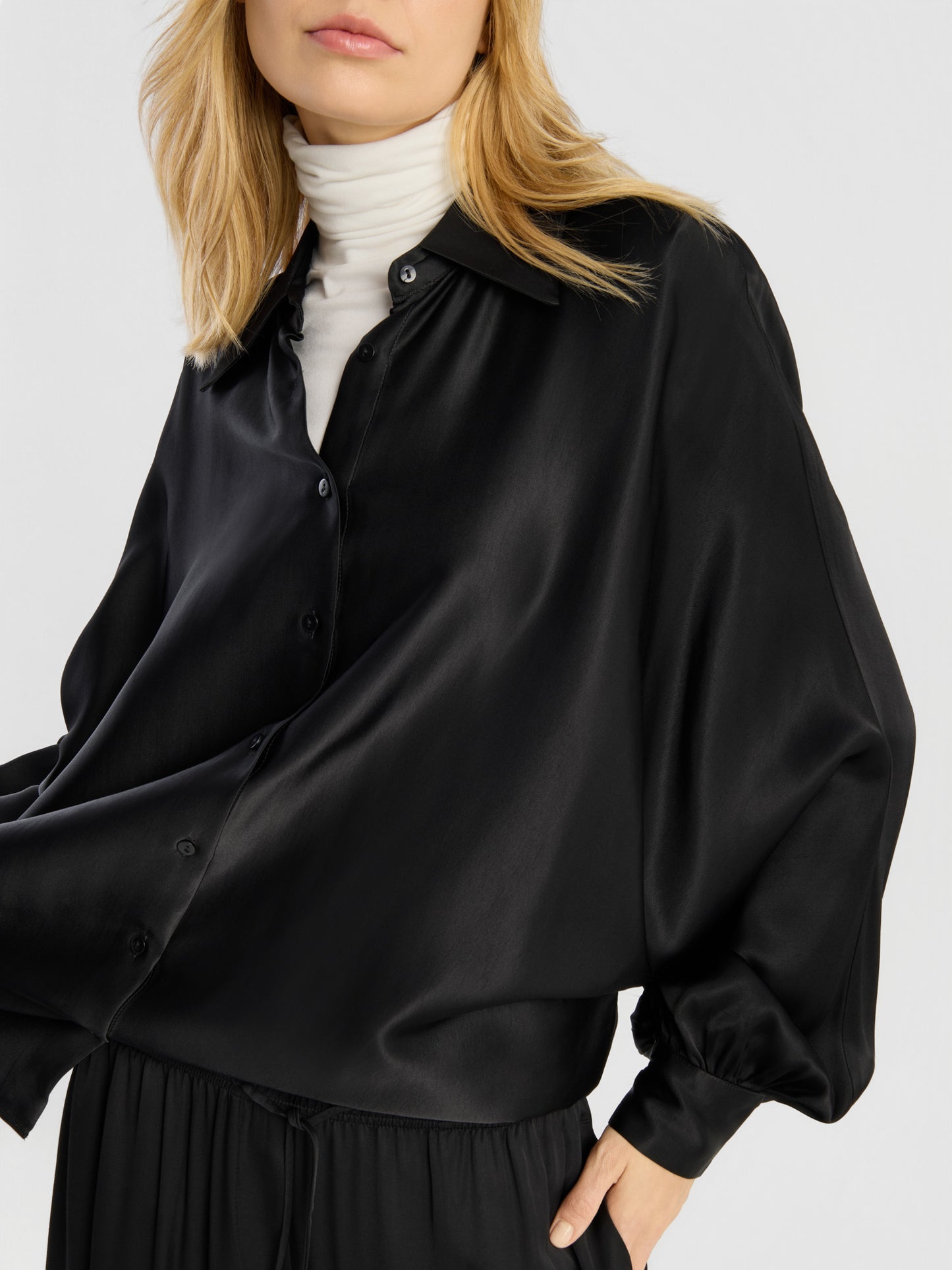 Eine Frau präsentiert die Bluse „Antonia Shiny Black One Size“ und trägt sie als Satinbluse mit Knöpfen über einem weißen Rollkragenpullover. Die Bluse hat weite Ärmel und wird zu einer schwarzen Hose kombiniert. Sie steht vor einem einfarbigen Hintergrund, wobei ihr Gesicht teilweise außerhalb des Bildausschnitts liegt.