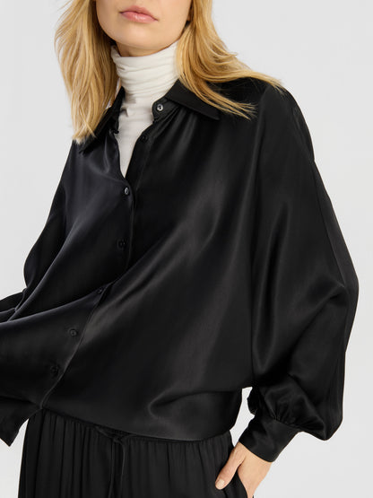 Eine Frau präsentiert die Bluse „Antonia Shiny Black One Size“ und trägt sie als Satinbluse mit Knöpfen über einem weißen Rollkragenpullover. Die Bluse hat weite Ärmel und wird zu einer schwarzen Hose kombiniert. Sie steht vor einem einfarbigen Hintergrund, wobei ihr Gesicht teilweise außerhalb des Bildausschnitts liegt.