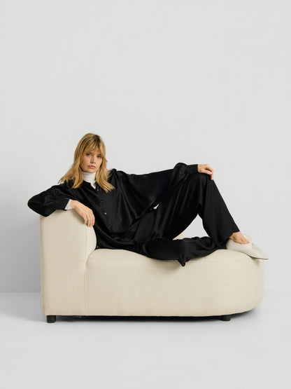 Eine Frau, die die Bluse „Antonia Shiny Black One Size“ trägt, sitzt auf einem cremefarbenen Sofa vor einem schlichten weißen Hintergrund und blickt mit entspannter Haltung und neutralem Gesichtsausdruck direkt in die Kamera.